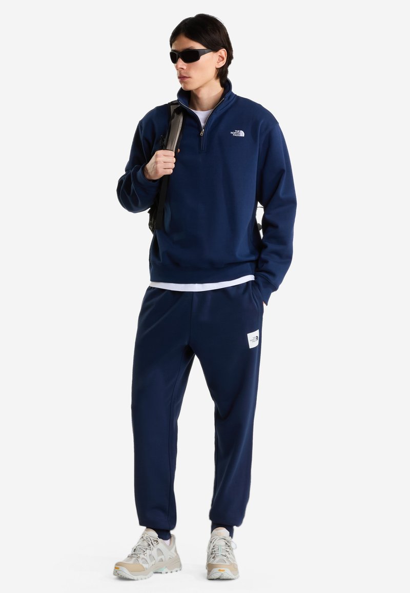 Hombre que lleva un chándal azul marino de North Face, zapatillas beige, gafas de sol negras y lleva una mochila al hombro.