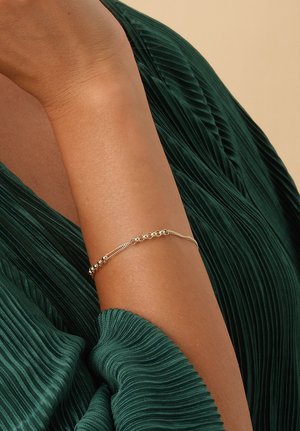 Arm wearing a delicate gold chain bracelet over a dark green pleated fabric background, showing a partial sleeve.