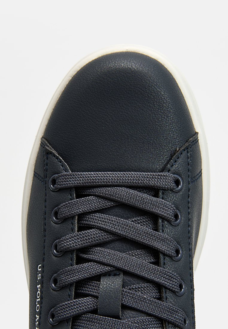 Baskets en cuir bleu marine foncé avec une capuche texturée, semelle en caoutchouc blanc rond et lacets gris. Marque "U.S. Polo Assn." sur le côté.