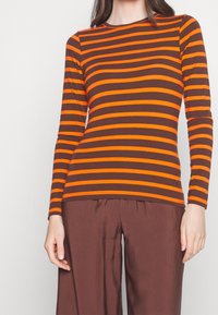 Langsleeve top in bruin met stevige oranje horizontale strepen, ronde halslijn en getailleerd ontwerp. Gecombineerd met losse bruine broeken.