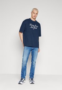 Camiseta de algodón azul marino con el logo "Tommy Jeans New York" en blanco, combinada con jeans azul desgastado y zapatillas deportivas multicolor.