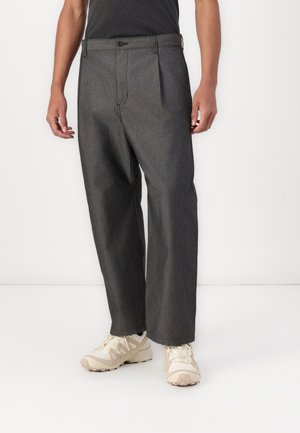 Mann trägt dunkelgraue weite Hose und beige Sneakers, steht vor schlichtem weißem Hintergrund.
