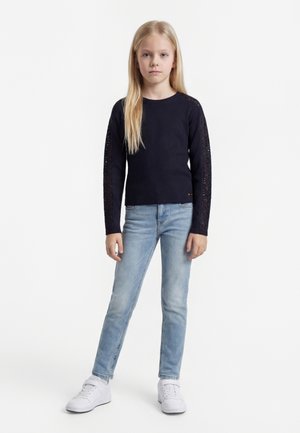 Mladé dievča s dlhými blond vlasmi, ktoré má na sebe čierny dlhý rukáv, svetlo modré rifle a biele tenisky, stojace pred bielym pozadím.