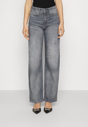 Vrouw die hooggetailleerde, wijdvallende grijze jeans draagt, gecombineerd met zwarte puntige hakken met zilveren strikaccenten.