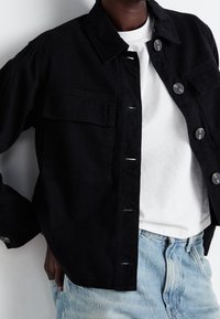 Musta farkkutakki, jossa on kaulus, kaksi etutaskua ja napit. Vaatteena valkoinen T-paita ja vaaleansiniset farkut.