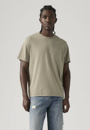 Mand med fletninger iført en beige T-shirt med lommer og slidte blå jeans, stående foran en ensfarvet lys baggrund.