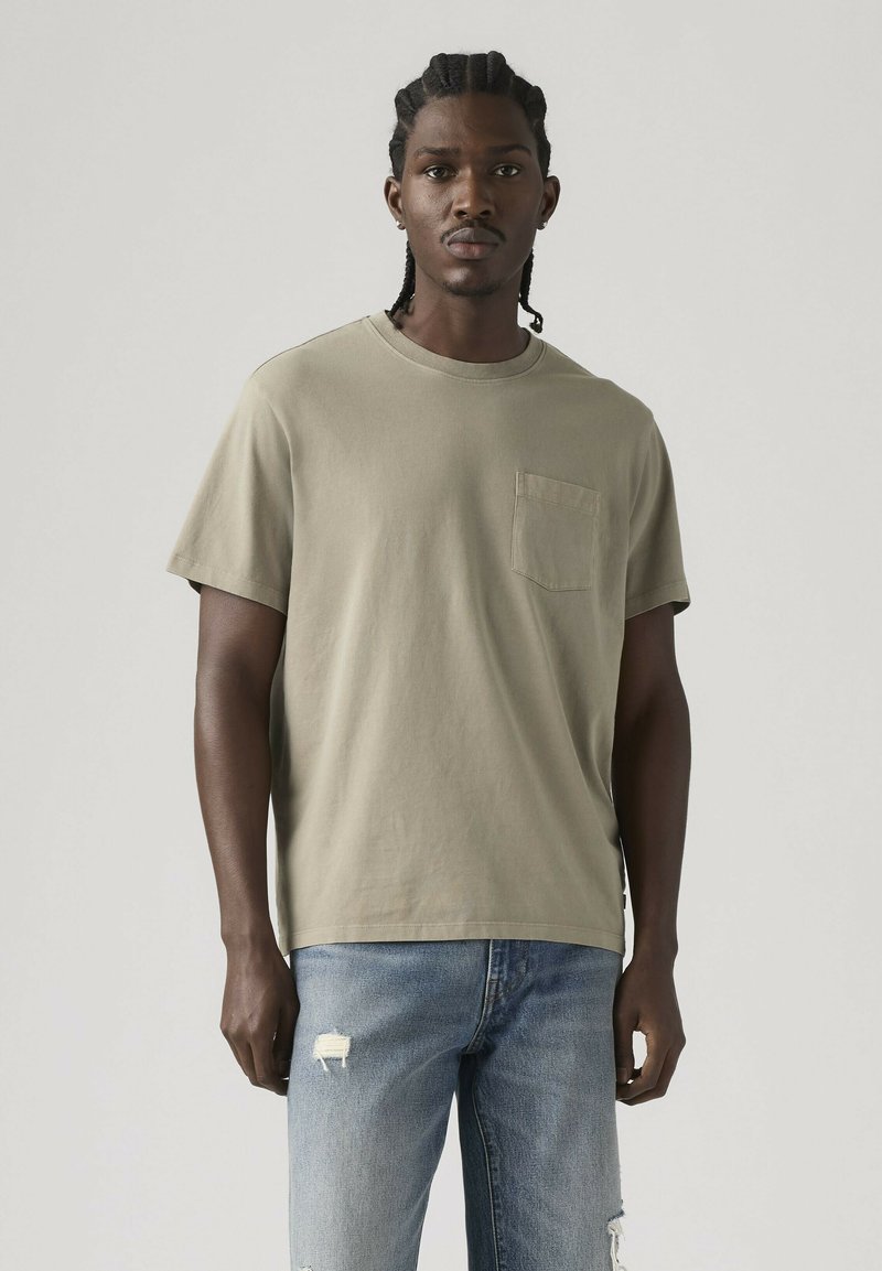 Homme aux cheveux tressés portant un t-shirt beige avec poche et un jean bleu déchiré, debout devant un fond clair uni.