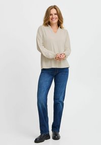 Beige, langærmet bluse med V-hals og rynkede manchetter, parret med blå denimjeans og sorte ankelstøvler, poseret mod en neutral baggrund.