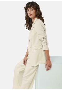 Beżowa dopasowana marynarka z pojedynczym guzikiem, klapami w kształcie litery V i wyraźnymi ramionami. W komplecie z szerokimi spodniami.