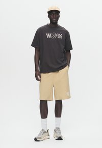 Graphic t-shirt in dark gray with logo print, beige shorts, and cream sneakers with textured accents. Accessories include a beige cap.