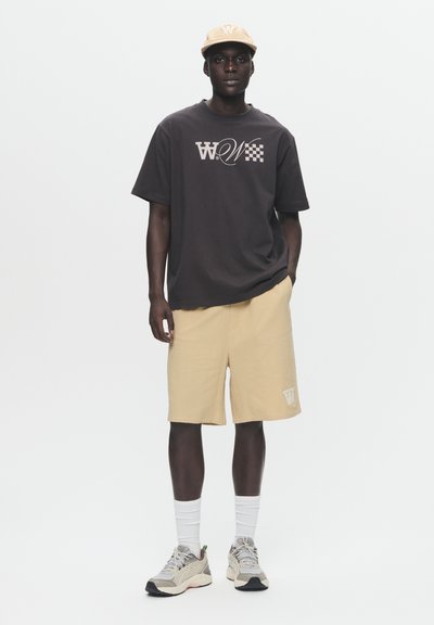 Grafisch T-shirt in donkergrijs met logo-print, beige shorts en crèmekleurige sneakers met textuuraccenten. Accessoires zijn een beige pet.