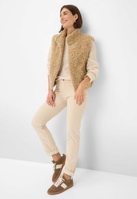 Eine Frau lächelt, trägt eine beigen flauschigen Weste über einem hellen Pullover, beige Hosen und braune Sneaker und steht vor einer weißen Wand.