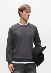 Szara bluza z ribbowanymi mankietami i dołem, z subtelnym czarnym logo. Model trzyma czarną czapkę, na jasnym tle.