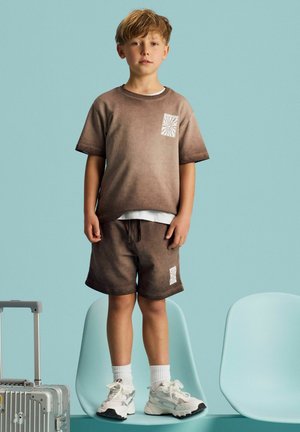 Young boy stands on light blue chair wearing brown gradient T-shirt and shorts with white sneakers, next to silver suitcase.