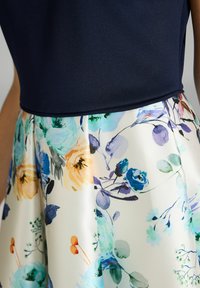 Robe florale avec un haut bleu marine et une jupe florale multicolore dans des teintes de bleu, pêche et vert sur un fond crème clair.