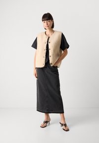 Beige Kunstfellweste mit Knopfverschluss, kombiniert mit einem schwarzen kurzärmeligen Oberteil und einem schwarzen Jeansrock, abgerundet mit braunen Absatzsandalen.