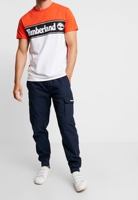 Muže v oranžovém a bílém tričku Timberland, navy cargo kalhotách a bílých teniskách, který stojí s jednou rukou v kapse.