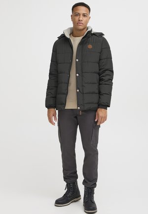 Chaqueta acolchada negra con capucha forrada de forro polar gris, botones a presión y parche de logo marrón, combinada con pantalones cargo oscuros y botas de cordones negras.