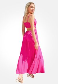 Robe maxi rose avec bretelles spaghetti ajustables, corsage froncé, jupe à volants et tissu fluide. Portée avec des sandales à lanières neutres.