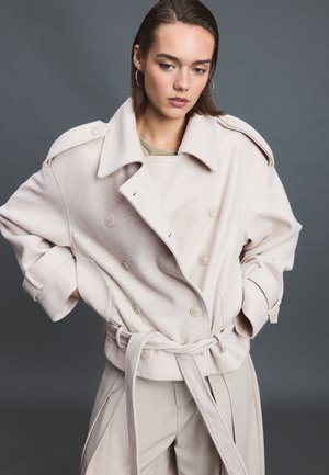 Jeune femme aux cheveux courts bruns portant des boucles d'oreilles créoles, un manteau croisé crème ceinturé, et un pantalon beige clair sur fond gris.