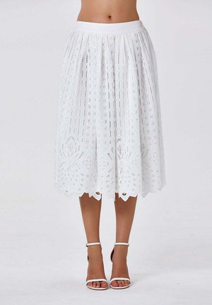 Jupe en dentelle blanche longueur genou avec motifs floraux et géométriques portée avec des talons blancs à bride cheville sur fond neutre.