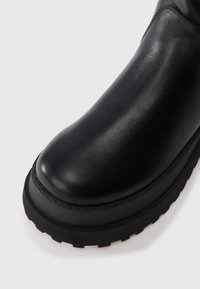 Bota de tornozelo em couro preto com bico arredondado, sola de borracha preta grossa e textura suave. A bota tem detalhes mínimos e um perfil elegante.