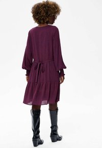 Paarse jurk met een patroon, lange pofmouwen, een tailleband en een uitlopende zoom. Draag met kniehoge zwarte leren laarzen met hak.