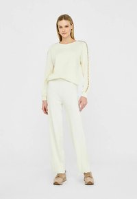 Svijetlo kremasti pleteni džemper s dugim rukavima, s detaljima sa strane. Uparen s odgovarajućim širokim hlačama. Model nosi bež tenisice.