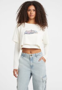 Crème cropped t-shirt met een grafisch ontwerp met een ster en tekst. Draagt over een lange mouw witte top, gecombineerd met lichtblauwe baggy jeans.
