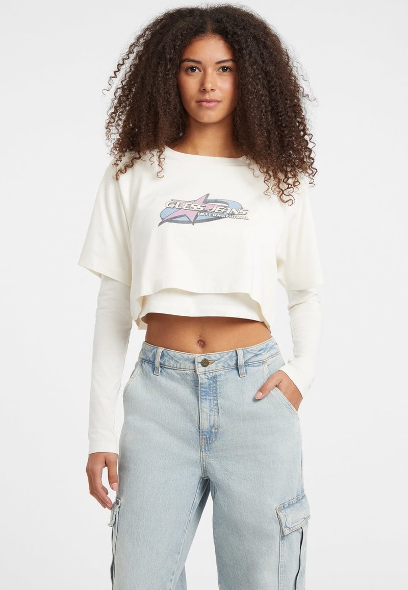 Crème cropped t-shirt met een grafisch ontwerp met een ster en tekst. Draagt over een lange mouw witte top, gecombineerd met lichtblauwe baggy jeans.