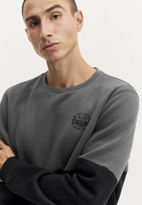 Grijze sweatshirt met een zwart onderste gedeelte, ronde halslijn en een klein gedrukt logo op de borst met "Built in Blue, Blend 1993."