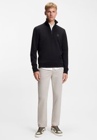 Schwarzes Sweatshirt mit Reißverschluss und Kragen, getragen über einem weißen Hemd. Helle, beige Hose und grüne, schwarze und weiße Sneaker komplettieren das Outfit.