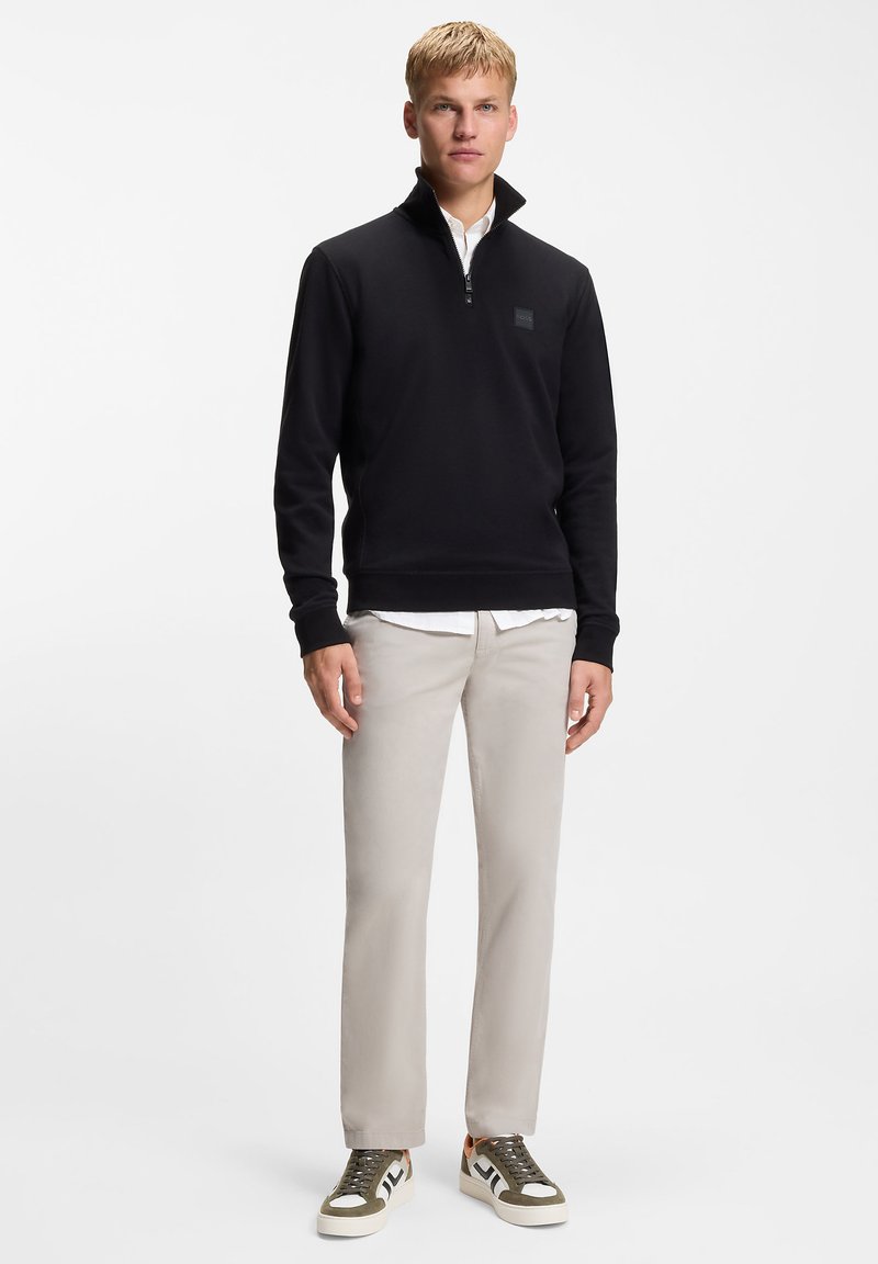 Schwarzes Sweatshirt mit Reißverschluss und Kragen, getragen über einem weißen Hemd. Helle, beige Hose und grüne, schwarze und weiße Sneaker komplettieren das Outfit.