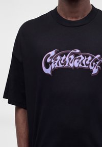 Κοντινό πλάνο ενός ατόμου που φοράει μαύρο T-shirt με μωβ και ροζ στυλιζαρισμένο κείμενο στο στήθος.