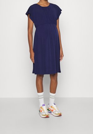 Zwangere persoon die een marineblauwe jurk tot op de knie draagt, witte kousen met grijze strepen en witte sneakers met oranje en groene accenten.