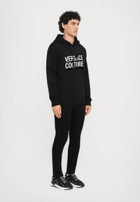 Čierna mikina s kapucňou a grafikou "VERSACE JEANS COUTURE". Slušivé čierne nohavice a čierne tenisky s bielymi akcentmi. Štruktúrovaná tkanina, neformálny dizajn.