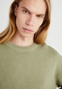 Pulover tricotat de culoare verde deschis, cu guler rotund, având un material texturat și guler cu nervuri. Vedere de aproape care pune accent pe detaliile îmbrăcămintei.
