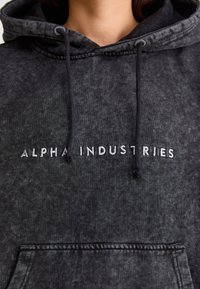 Juodas džemperis su tekstūru, su gaubtu ir raišteliais. Priekyje yra balta siuvinėta "ALPHA INDUSTRIES" užrašas.