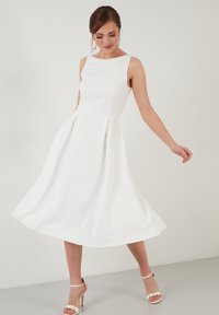 Rochie albă fără mâneci, cu fustă evazată, realizată dintr-un material neted, cu guler rotund și plii subtile. Accesorizată cu pantofi cu barete.