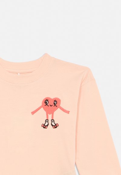 Hellrosa langärmliges Hemd mit einem lächelnden Herz-Charakter, der weiße Socken und rosa Rollschuhe trägt, auf der Brust gestickt.