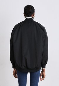 Čierna bomber bunda s rebrovaným golierom, elastickými manžetami a pásom. Hladká, ľahká tkanina s voľným strihom a jemnými bielymi akcentmi.