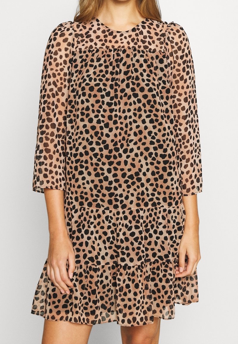 Femme portant une robe marron longueur genou avec des taches noires irrégulières, des manches 3/4 transparentes et un ourlet à volants superposés.