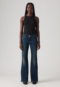 Nainen, jolla on pitkät letit, pukeutunut mustaan hihattomaan puseroon, tummansiniset leveälahkeiset farkut ja mustat kärjetön kengät, seisoo vaaleaa taustaa vasten.