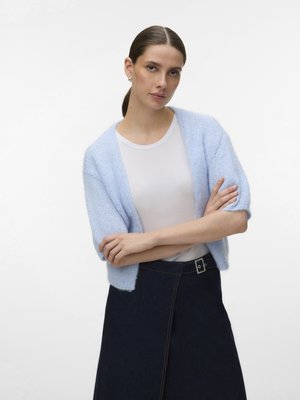 Jeune femme aux cheveux bruns en queue de cheval, portant un cardigan tricoté bleu clair sur un t-shirt blanc et une jupe en denim foncé, bras croisés.