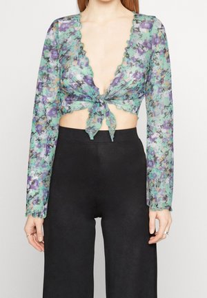 Hun har en gennemsigtig, langærmet blomstret cropped top med bindebånd foran og højtaljede sorte bukser på, stående mod en ensartet baggrund.