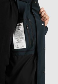 Meremehe sisu sisaldab tõmblukuga taskut ja hooldusjuhiseid sisaldavat silti. Kanga pind on sile ja tume, mati viimistlusega.