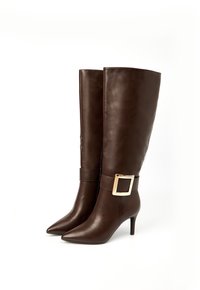 Bottes en cuir brun montant jusqu'au genou avec des pointes, des talons hauts et un détail de boucle carrée en or à la cheville. Texture lisse, design élégant.