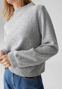 Donna che indossa un maglione lavorato a maglia grigio testurizzato con maniche lunghe abbinato a jeans blu, in piedi contro uno sfondo chiaro e uniforme.