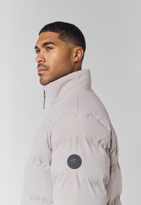 Jachetă puffer de culoare gri deschis cu guler înalt, având o etichetă logo rotundă pe mâneca stângă și o textură de țesătură moale și netedă.