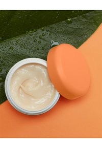 Witte pot met een romige, lichtbeige inhoud met een gestructureerd oppervlak, afgewerkt met een gladde matte oranje deksel, tegen een achtergrond van oranje en groen.